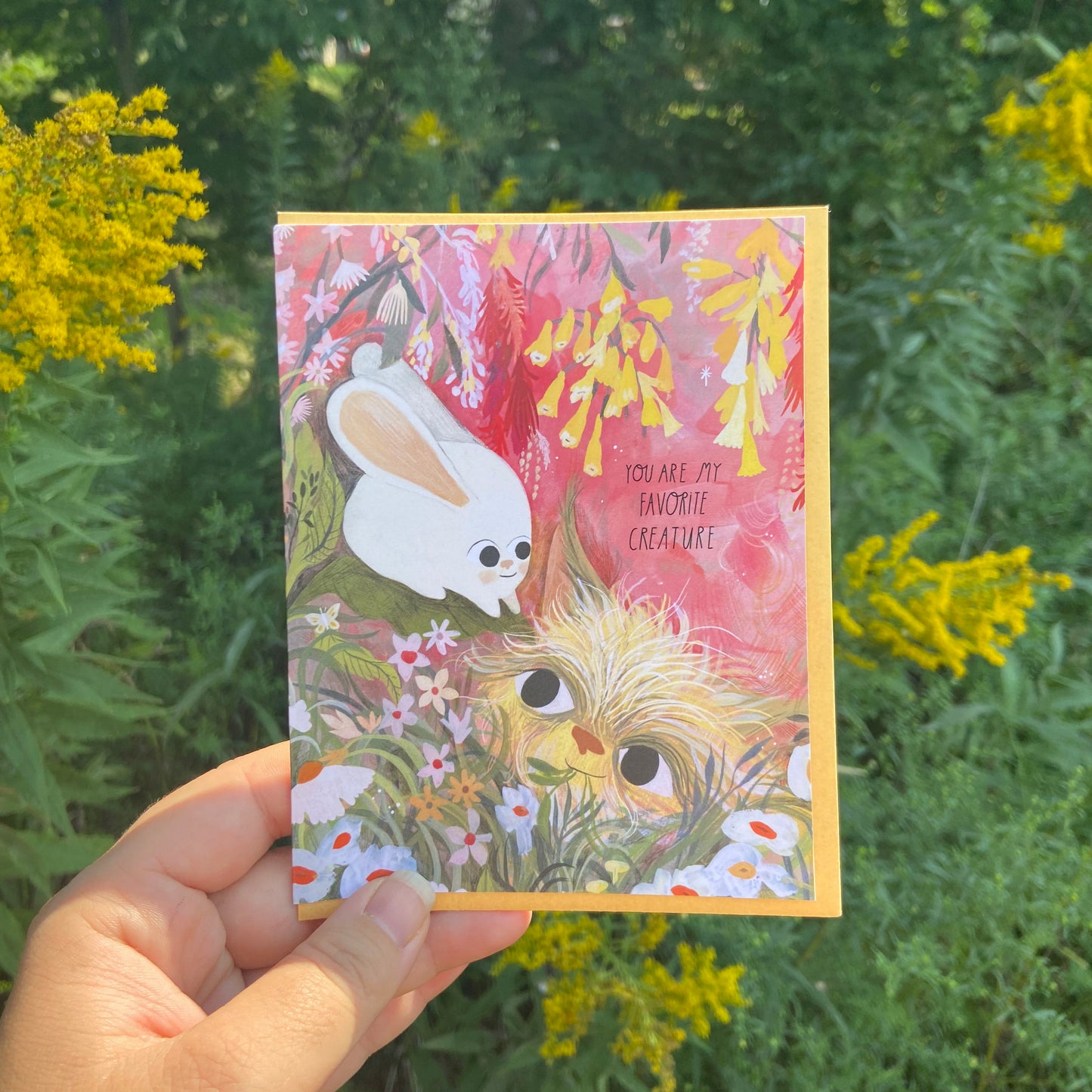 Greeting card titled ‘You Are My Favorite Creature’ featuring a whimsical illustration of a furry creature and a small white bunny in a flower-strewn field under a red sky, 4.25″×5.5″ folded card with Kraft envelope.